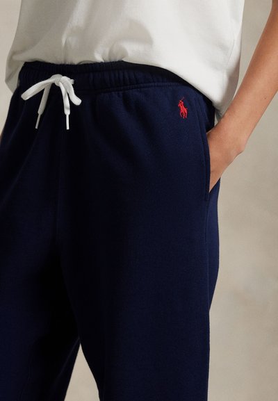 Άτομο με το δεξί χέρι στην τσέπη, φορώντας μπλε σκούρες φόρμες με λευκό κορδόνι και κόκκινο κεντημένο λογότυπο, συνδυασμένες με λευκό πουκάμισο.