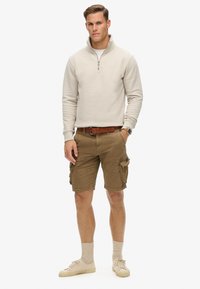 Pull en polaire zippé beige clair, shorts cargo marron avec plusieurs poches, ceinture en cuir marron, chaussettes crème et baskets beiges.