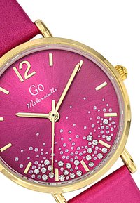 Montre rose avec un boîtier doré ; le cadran rond est orné de pierres précieuses scintillantes, avec des index en or et des aiguilles, sur un bracelet en rose doux.