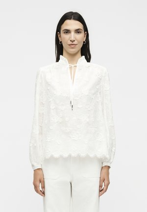 Γυναίκα με μακριά σκούρα μαλλιά που φοράει λευκό δαντελωτό μπλουζάκι με λουλουδένια σχέδια και λεπτομέρεια δέσιμου στον λαιμό, συνδυασμένο με λευκό παντελόνι, κοιτάζει προς τα εμπρός.