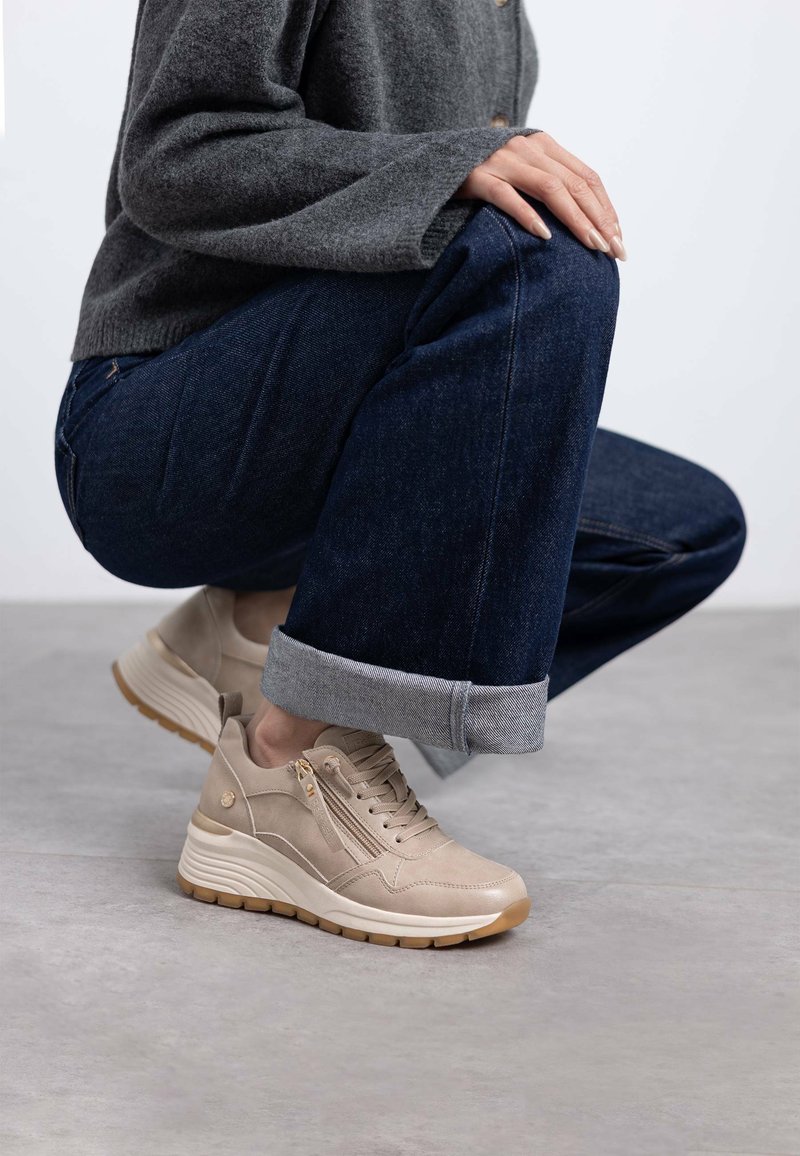 Zapatillas beige con parte superior de ante texturizado, que cuentan con un bolsillo lateral con cremallera, cuello acolchado y suela de goma, combinadas con jeans oscuros.