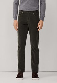 Man in donkergroene corduroy broek, grijze lange mouwen trui en donkerbruine sneakers met witte zolen, staand tegen een effen achtergrond.