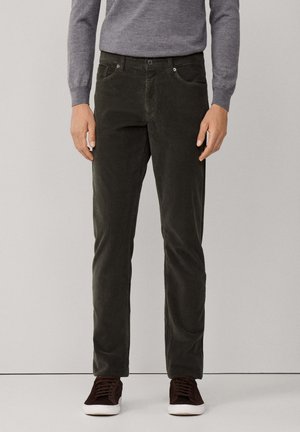Uomo che indossa pantaloni in corda verde scuro, un maglione grigio a maniche lunghe e scarpe da ginnastica marroni scure con suole bianche, in piedi davanti a uno sfondo semplice.