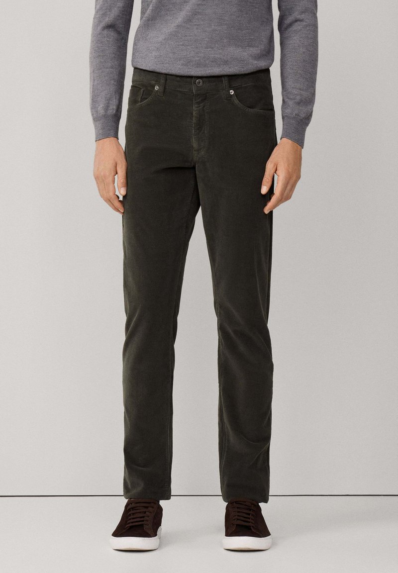 Man in donkergroene corduroy broek, grijze lange mouwen trui en donkerbruine sneakers met witte zolen, staand tegen een effen achtergrond.