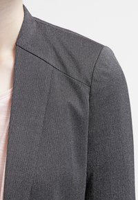Blazer grigio con tessuto testurizzato, caratterizzato da un colletto netto e linee pulite, parzialmente visibile insieme a una maglietta rosa chiaro sottostante.
