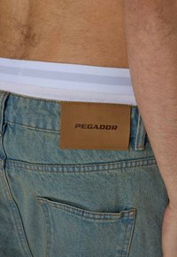 Light blue denim jeans with a brown leather patch featuring the text "PEGADOR." The jeans have a classic cut and visible stitching details.