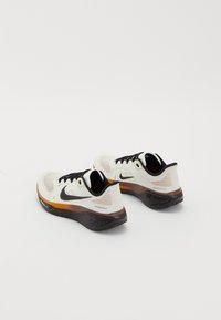 Paire de chaussures de running blanches Nike avec un logo noir, des lacets noirs et des semelles tachetées orange et noir sur fond blanc.