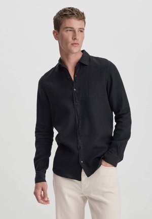 Chemise en lin noir avec col boutonné et manches longues, présentant une coupe décontractée et une texture lisse, associée à un pantalon beige clair.