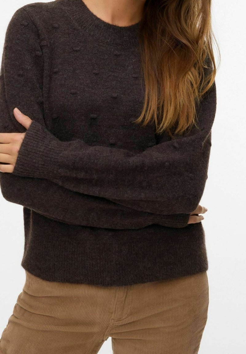 Femme portant un pull texturé marron foncé à manches longues, les bras croisés sur un pantalon en velours côtelé couleur beige, sur fond blanc.