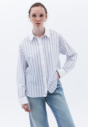 Camisa blanca de botones con rayas verticales azules, de manga larga y con cuello puntiagudo. Combinada con jeans de mezclilla de color azul claro.