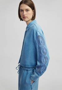 Chaqueta de mezclilla azul claro con cuello y mangas largas, adornada con intrincados detalles de ganchillo azul. Cuerda reguladora en la cintura.