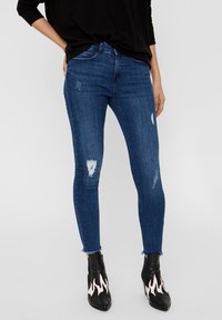 Vaqueros ajustados de color azul con parches desgastados y dobladillos deshilachados, hechos de tela de mezclilla. Combinados con botas negras que tienen detalles de llamas.