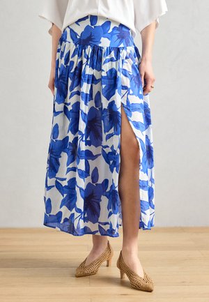 Blue floral print maxi skirt with a side slit, featuring a fitted waistband and flowing fabric. Worn with beige, patterned high-heeled shoes.