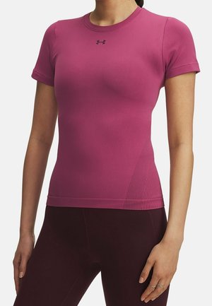 Kvinde iført en tætsiddende magenta sportsskjorte med korte ærmer og mørke træningsleggings, stående med den ene hånd afslappet ved siden af sig.