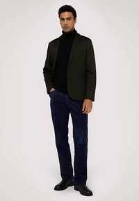 Mörkgrön kavaj med hakkrage, svart polotröja och blå velourbyxor. Modellen bär svarta läderstövlar och står i en pose mot en ljus bakgrund.