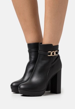 Bottes noires à la cheville en cuir lisse, avec un talon haut épais, un bout rond et une décoration en chaîne dorée autour de la cheville.