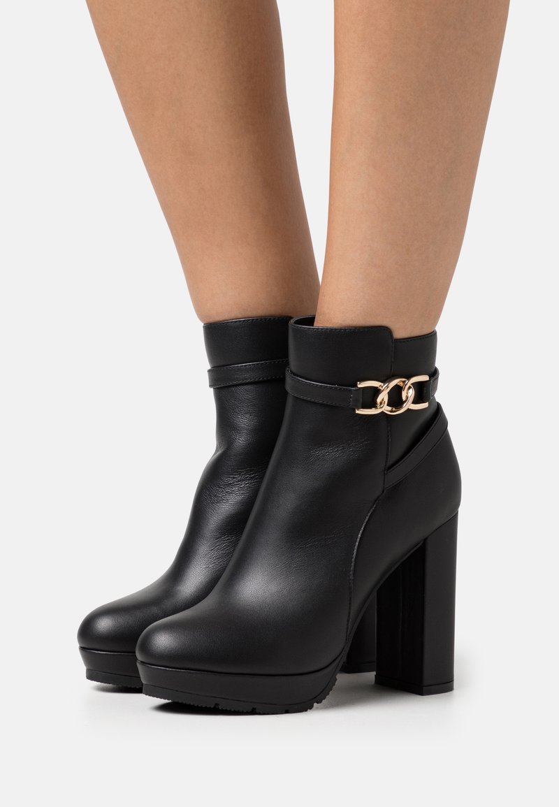 Botas negras de tobillo hechas de cuero liso, con un tacón alto y grueso, puntera redonda y un acento de cadena dorada alrededor del tobillo.