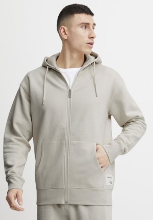 Light gray zip-up hoodie made of soft fabric with a kangaroo pocket, drawstring hood, and ribbed cuffs. Features a small logo patch.