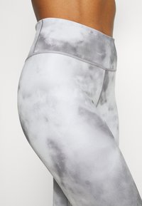 Close-up van een persoon die grijze en witte tie-dye leggings met hoge taille draagt, met zicht op de zijkant van de taille en heup tegen een effen achtergrond.