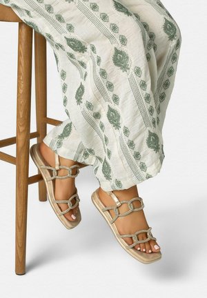 Jambes croisées sur un tabouret en bois portant un pantalon blanc à motifs floraux verts et des sandales plates dorées avec des brides à maillons de chaîne.