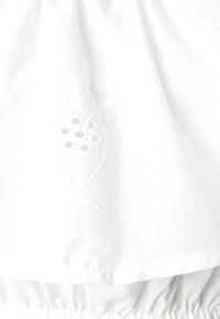 Tissu en coton blanc avec broderies florales présentant un motif circulaire. L'ourlet est froncé, créant une apparence texturée et douce.