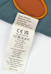 Tag z bavlněného polštáře, béžový s černým textem. Uvedeny pokyny pro údržbu; materiál 100% organická bavlna, výplň z polyuretanové pěny. Vyrobeno v Indii.