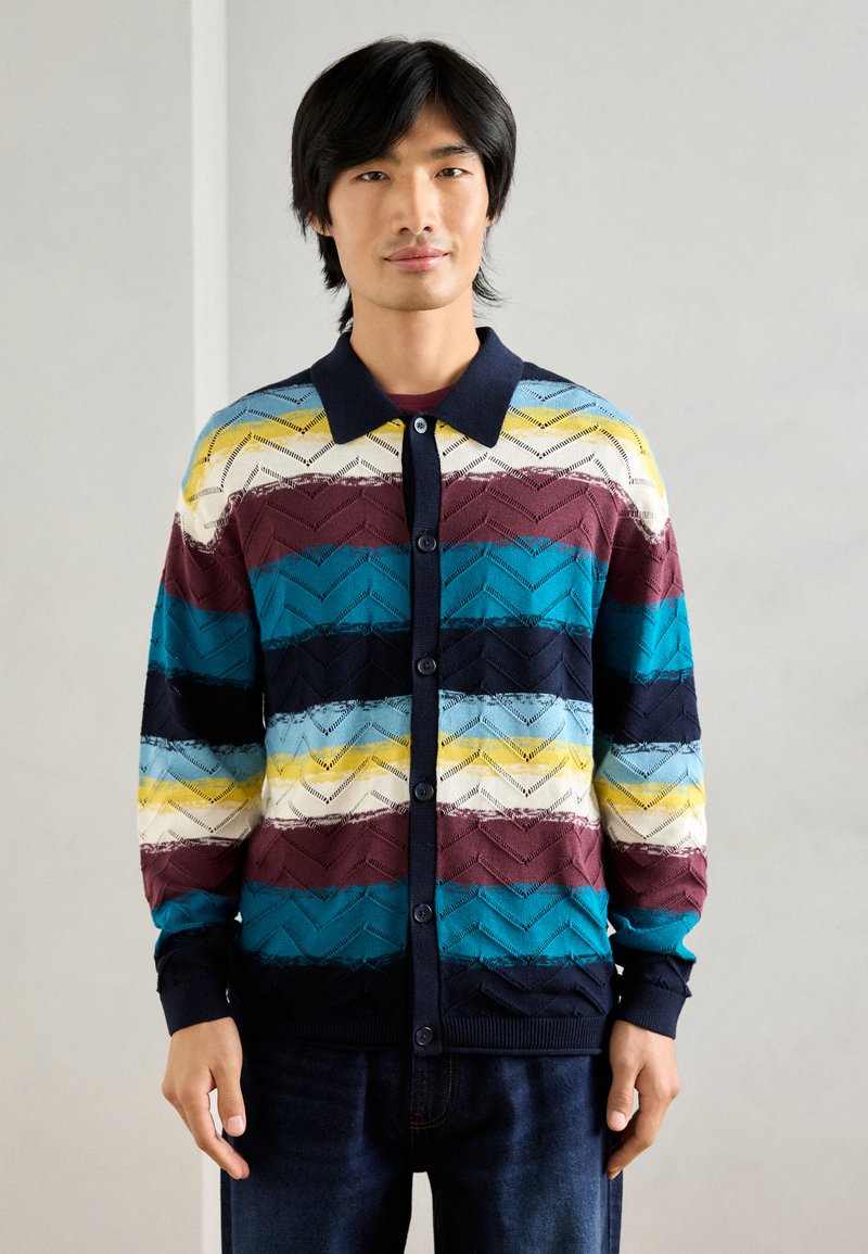 Cardigan tricoté multicolore avec un motif en chevrons, arborant un col et des boutons bleu marine, dans des teintes de teal, bourgogne, jaune et blanc.