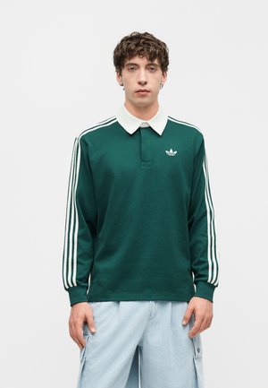 Młody mężczyzna z kręconymi włosami, ubrany w zieloną, długorękawową koszulkę Adidas z białymi paskami i kołnierzykiem, do tego jasnoniebieskie spodnie typu cargo.