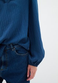 Kék, textúrált blúz hosszú, puffos ujjakkal, sötét farmernadrággal, klasszikus fazonban. Finom gyűrődés minta díszíti.