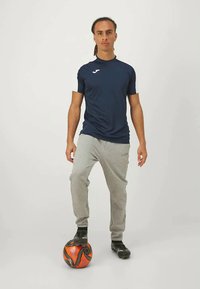 Camiseta atlética azul-marinho, tecido leve, mangas curtas, combinada com calças de moletom cinza-claro, chuteiras pretas, em pé sobre uma bola de futebol laranja e preta.