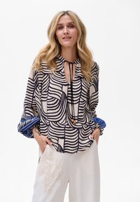 Losse blouse in beige met zwarte abstracte lijnen, voorzien van een keyhole-halslijn en een trekkoord. De manchet heeft blauwe patroonaccenten.