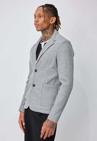 Homme avec des tatouages sur le cou et les mains, portant un blazer gris clair avec des boutons et des poches avant, debout devant un fond clair uni.