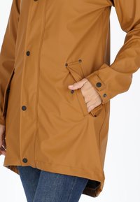 Giacca impermeabile marrone con chiusura a bottoni a pressione, tasche laterali e una texture liscia con dettagli neri.