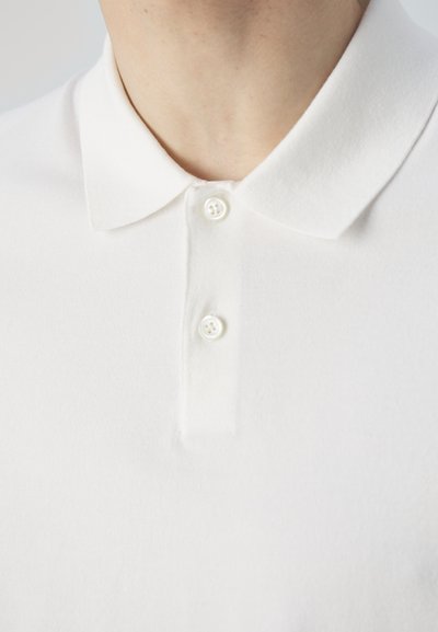 Hvit polo skjorte med en myk tekstur, med klassisk krage og tre knappeåpning. Knapper er runde og stoffet er glatt.