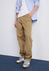 Tan, relaxed-fit pants with a smooth texture, paired with silver sneakers. Worn with a white shirt and blue striped overshirt.