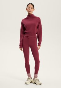 Set sportiv bordeaux compus dintr-un hanorac cu guler înalt și colanți ajustați. Materialul este neted, cu manșete zimțate și șosete asortate. Pantofi neutri.