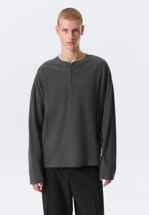 Jeune homme aux cheveux blonds courts portant un t-shirt henley à manches longues gris foncé ample et un pantalon noir sur un fond clair uni.