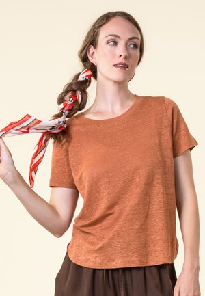 Frau in einem rostfarbenen T-Shirt mit kurzen Ärmeln, die einen rot-weiß gestreiften Schal in ihrem Haar geflochten hält und zur Seite blickt.