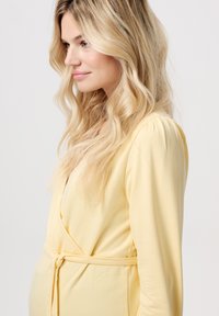 Geel wikkeljurk met lange mouwen, gemaakt van zachte stof. Heeft een riem bij de taille en lichtelijk volumineuze schoudernaden. Soepele textuur.