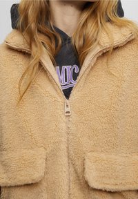 Veste zippée en fausse fourrure beige avec de grandes poches plaquées, tissu texturé et col haut, superposée à un sweat à capuche sombre.