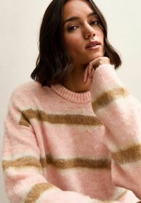 Roze wollen trui met bruine strepen, voorzien van een geribde halslijn en een zachte, fuzzy textuur. Model poseert met de hand op de kin.