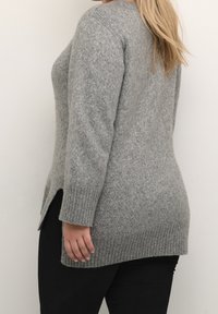 Grå strikket sweater med ribbede manchetter og afslutning, der har sidesplitter. Texturen virker blød og hyggelig. Den er parret med sorte bukser.