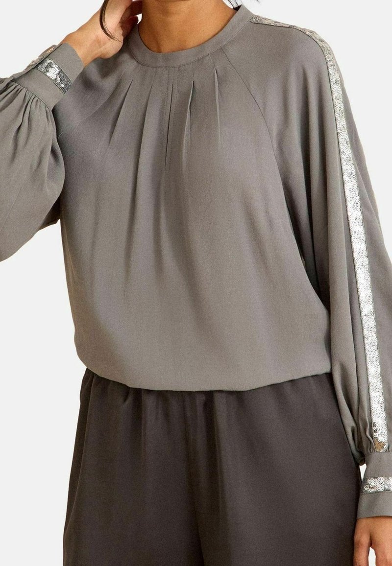 Femme portant un chemisier gris ample avec une bande de sequins argentés le long des manches et un pantalon gris assorti, main touchant le cou.
