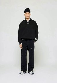 Man wearing a black beanie, oversized black collared sweatshirt, loose black pants, and white sneakers, standing against a plain light background.