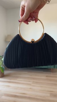 Schwarze gefaltete Handtasche mit einem goldenen runden Griff und einem kugelförmigen Verschluss. Glatte Textur und geschwungene Form.