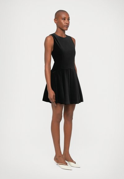 Zwarte halterjurk van fluwelen stof met een nauwsluitende bovenkant en een uitlopende rok. Gecombineerd met witte instappers, weergegeven tegen een effen achtergrond.