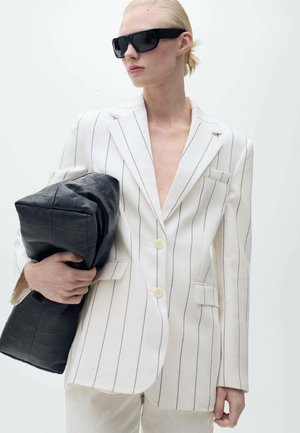 White pinstriped blazer featuring a deep V-neck, two buttons, and side pockets, paired with a black quilted bag held in one hand.