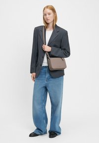 Borsa a tracolla marrone con superficie liscia, chiusura con zip e tracolla regolabile. Indossata da una persona con un blazer grigio aderente e jeans a gamba larga.