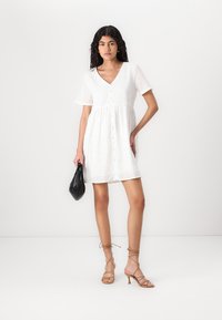 Bílá bavlněná šaty s krátkými rukávy, výstřihem do V, předním zapínáním na knoflíky a vzorem s očky. Doplněné béžovými sandály na podpatku a černou kabelkou.