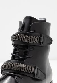 Botín negro de cuero hasta el tobillo con punta redonda, que presenta dos correas decorativas adornadas con strass y cordones negros. Textura suave con acentos metálicos.